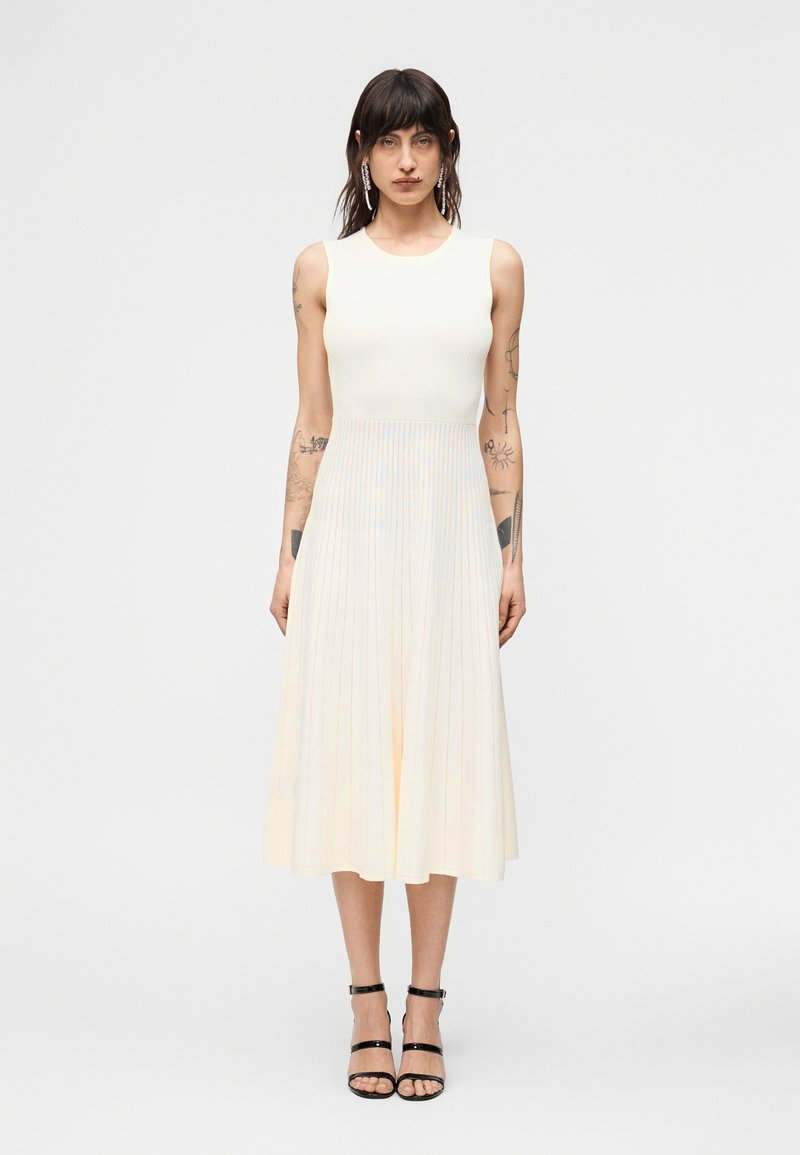 Γυναίκα με σκούρα μαλλιά και τατουάζ που φοράει  αμάνικο, κρεμ πλισέ μεσοφόρεμα και μαύρα πέδιλα με λουράκια, στέκεται μπροστά σε απλό φόντο.