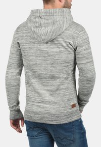 Gray marled hoodie with a textured finish, drawstring hood, and a small brown leather logo patch at the lower side. Worn with blue jeans.