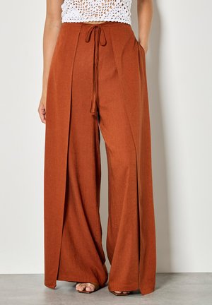 Weit geschnittene rostorangefarbene Hosen mit vorderen Falten, einem Kordelzug in der Taille und seitlichen Taschen, kombiniert mit einem weißen gehäkelten, kurz geschnittenen Oberteil.
