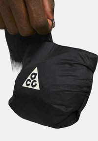 Svart nylonpåse med en triangelformad vit logga, hålls av en hand. Texturen är slät, med en kompakt och lätt design för portabilitet.