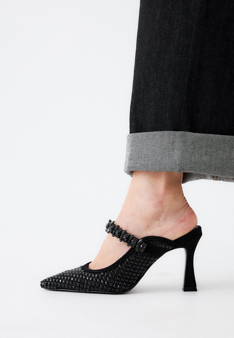 Mulas negras de tacón alto con punta afilada, textura tejida y correa decorativa en el tobillo con piedras preciosas negras. Jeans remangados parcialmente visibles.