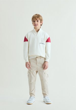 Blondýnský chlapec v bílém pulovru s červenými akcenty, béžových cargo kalhotách a bílých teniskách, stojící před jednoduchým světlým pozadím.