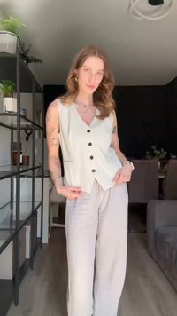 Off-white mouwloos vest met vijf zwarte knopen, gecombineerd met lichte grijze wijdpijpen broek, tegen een donkere achtergrond. Natuurlijk licht benadrukt de texturen.