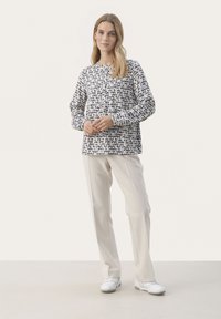 Langærmet, mønstret bluse i hvid med marineblå og sorte abstrakte former, parret med lyse beige bukser og hvide sneakers.