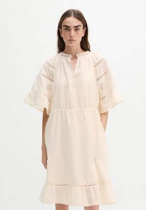 Vrouw met lang bruin haar draagt een losse, licht crèmekleurige jurk met brede kanten mouwen en een gerimpelde zoom, staand tegen een effen achtergrond.