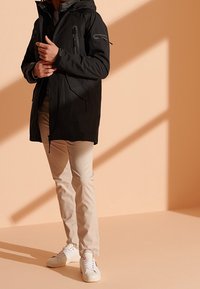 Chaqueta impermeable negra con capucha, bolsillos con cremallera y dobladillo ajustable; combinada con pantalones beige y zapatillas blancas sobre un fondo color durazno.