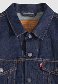 Kurtka jeansowa w ciemnym niebieskim kolorze z kontrastującym pomarańczowym przeszyciem, metalowymi guzikami i dwoma przednimi kieszeniami. Posiada klasyczny kołnierz i metkę z logo.