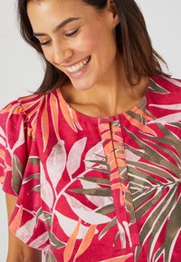 Blusa roja y rosa con un estampado floral, que presenta hojas verdes y blancas, un escote redondo y detalles de pliegues sutiles en el frente.