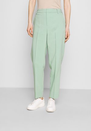 Persona che indossa pantaloni sartoriali a vita alta verde salvia e sneakers bianche, in piedi davanti a uno sfondo semplice di colore grigio chiaro.