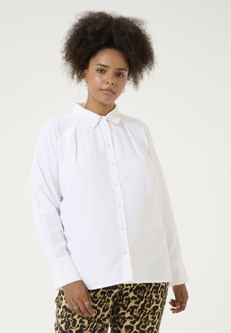 Chemise blanche à boutons, fabriquée dans un tissu lisse, avec un col classique et des manches longues. Design notable de manches raglan et coupe décontractée.