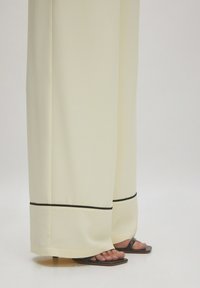 Pantalons larges crème avec un bord noir à l'ourlet, en tissu lisse. Associés à des sandales plates noires, mettant en avant des pieds nus.
