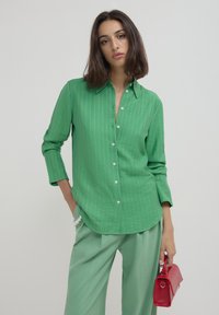 Woman wearing a green striped button-up shirt and light green pants, holding a small red handbag with a pearl bracelet on her wrist.