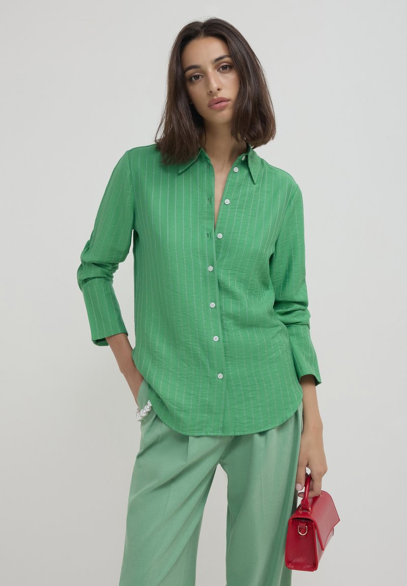 Woman wearing a green striped button-up shirt and light green pants, holding a small red handbag with a pearl bracelet on her wrist.