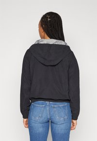 Svart hettejakke med stripete innerfôr, avslappet passform og elastiske mansjetter; paret med lyseblå denimjeans og en teksturert flettefrisyre.