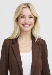 Blondínka sa usmieva, má na sebe hnedé sako cez biely top, stojí pred jednoduchým svetlosivým pozadím.