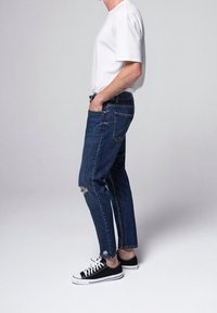 Person wearing white t-shirt, ripped blue jeans, and black sneakers, standing sideways with hands in pockets against plain background.