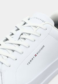 Gros plan d'une basket blanche Tommy Hilfiger avec des lacets blancs et le logo sur le côté et la languette sur un fond clair uni.