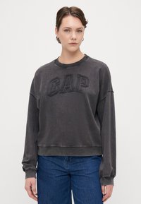 Sudadera gris corta con el logo texturizado de "GAP" en el frente, cuello redondo y mangas largas. Combinada con jeans de mezclilla azules.