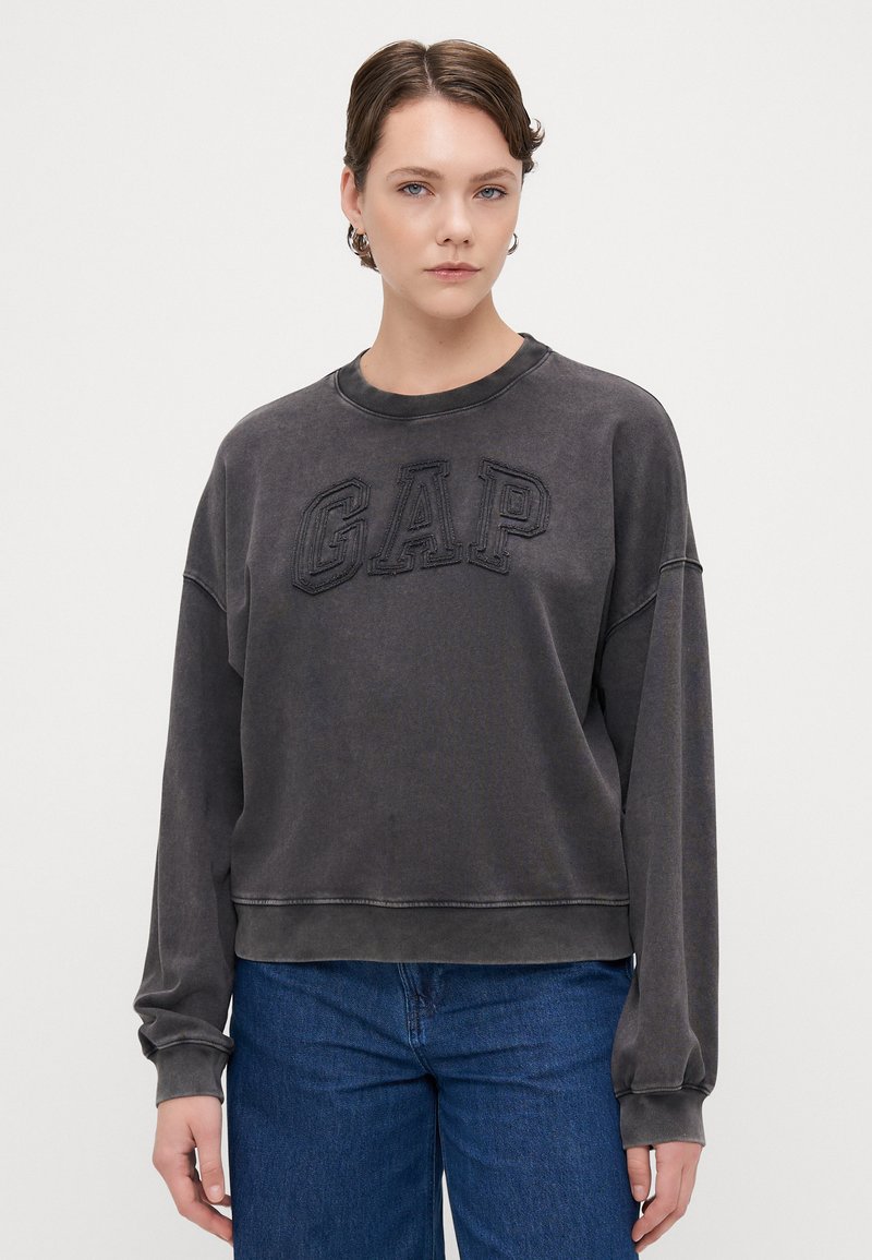 Sudadera gris corta con el logo texturizado de "GAP" en el frente, cuello redondo y mangas largas. Combinada con jeans de mezclilla azules.