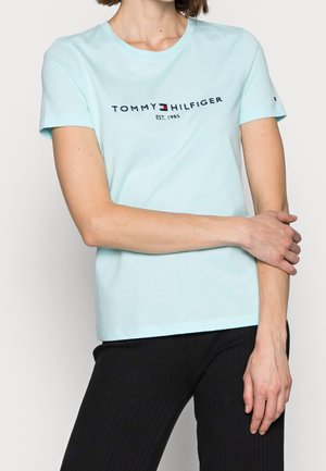Nainen, joka pukeutunut vaaleansiniseen Tommy Hilfiger -t-paitaan, jossa on logo ja teksti "EST. 1985", yhdistettynä mustiin ribbineulospanteljoihin, kädet ristissä vyötärön kohdalla.