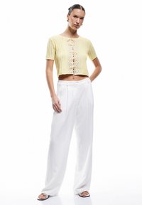 Gehaakte gele crop top met korte mouwen, voorzien van een voorpaneel met vetersluiting. Gecombineerd met witte wijde broek en zwarte sandalen.