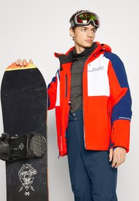 Helle rote und marineblaue Schneejacke mit weißen Akzenten, kombiniert mit dunkler Hose. Das Modell hält ein schwarzes Snowboard mit grafischem Design und Bindungen.