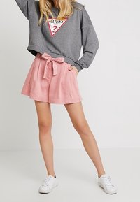 Harmaa huppari, jossa on kolmionmuotoinen logo, ja vaaleanpunaiset shortsit, joissa on rusetti vyötäröllä. Yhdistettynä valkoisiin lenkkikenkiin.