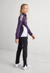 Violeta sporta jaka ar baltām detaļām un trim svītrām, kombinēta ar melnām biksēm, kurām ir teksturēts paneļis un rāvējslēdzēja detaļa pie potītes.