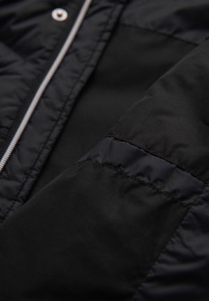 Chaqueta acolchada negra con textura acolchada, tela suave, cremallera plateada y botones a presión, con una vista en primer plano de la manga y las costuras.