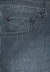 Jeans in denim di colore blu scuro con una leggera texture sbiadita, dotati di una piccola tasca e un dettaglio cucito a forma di "J" vicino alla tasca.