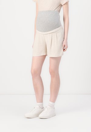 Schwangere Person trägt beige Umstandsshorts mit grauem Stretchbund, ein helles Oberteil, weiße Socken und weiße Turnschuhe vor weißem Hintergrund.