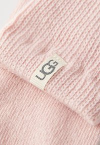 Tissu tricoté rose avec une texture douce ; comporte une étiquette blanche avec "UGG" en gris. Gros plan, montrant le détail du tissu et la couleur.