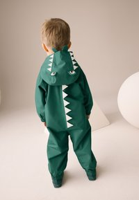 Grønt vandtæt regntøj med dinosaurmotiv, der har hætte, hvide trekantede detaljer og øjne på ryggen. Elastiske manchetter ved håndled og ankler.