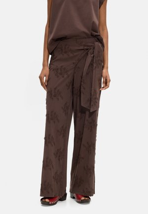 Kobieta nosząca brązowe haftowane szerokie spodnie z wysokim stanem z bocznym wiązaniem oraz czerwone sandały na platformie z odkrytymi palcami i ćwiekami.