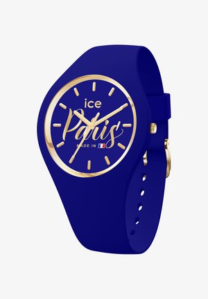 Reloj de silicona azul con esfera redonda, marcadores y manecillas de color dorado, y texto en cursiva "París". Presenta una correa con hebilla.