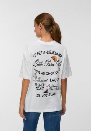 T-shirt blanc surdimensionné avec texte noir et graphiques de pâtisseries au dos, comprenant des phrases en français liées aux articles du petit-déjeuner.