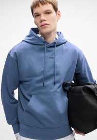 Giovane indossante un hoodie blu BOSS, che tiene una grande borsa di pelle nera contro uno sfondo bianco semplice.