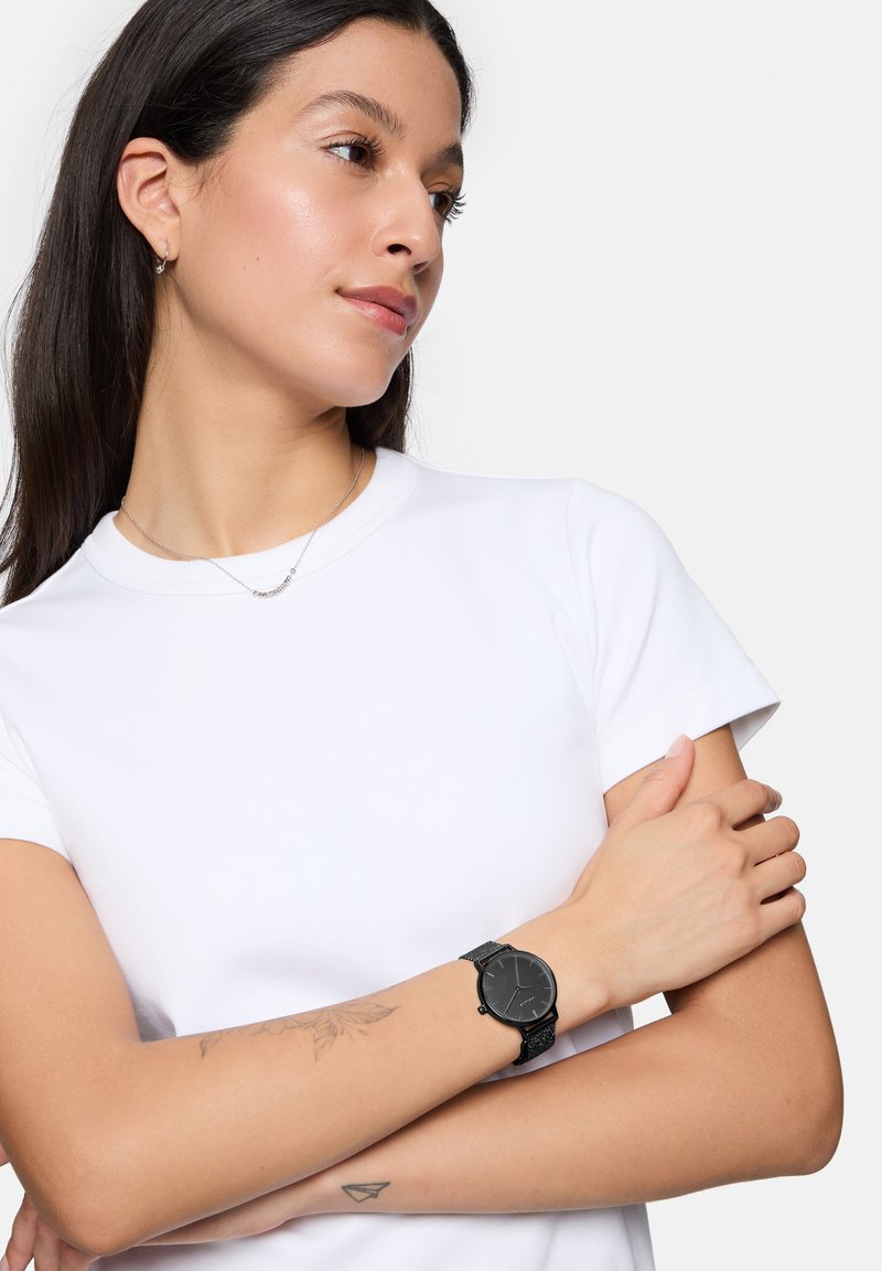 Schwarze Armbanduhr mit strukturiertem Armband und rundem Ziffernblatt, getragen mit einem schlichten weißen T-Shirt, das einen dezenten Tätowierung auf dem Arm zeigt.