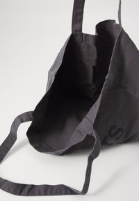 Borsa tote in cotone grigio scuro con manici lunghi, aperta e vuota, posata su una superficie bianca.