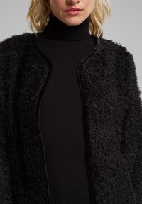 Svart, fluffig cardigan med en texturerad yta, öppen framsida och rund krage, bärs över en svart ribbad polotröja.