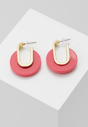 Prostokątne złote kolczyki-koła z dużymi błyszczącymi różowymi okrągłymi dyskami, przedstawione na białym sześcianie.