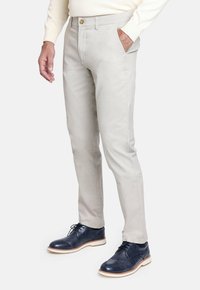 Hombre vestido con pantalones slim-fit de color beige claro, suéter crema y zapatos de vestir de cuero azul marino con suelas blancas, sobre un fondo blanco.