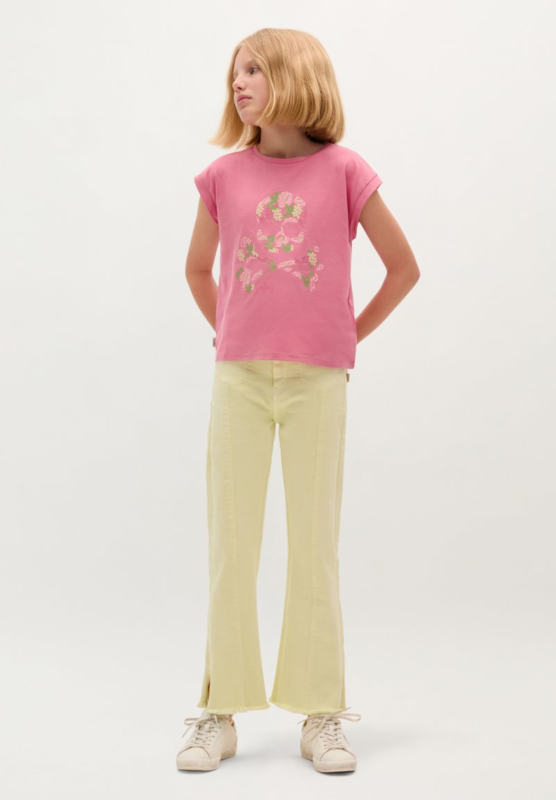 Roze T-shirt met korte mouwen en een bloemen- en schedelontwerp, gecombineerd met lichtgele flare jeans met zijslits. Witte sneakers maken de outfit compleet.
