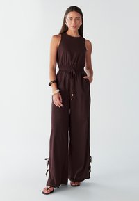 Brown sleeveless jumpsuit featuring an elastic waist, side pockets, and drawstring details at the ankles. Smooth fabric with a relaxed fit.