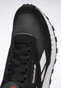 Svart sneaker med mesh- og semsket skinn-overdel, svarte snøringer, hvite detaljer, gummisåle, og røde detaljer på tåen. Reebok-logoen er synlig.