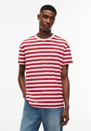 Hombre con camiseta de rayas horizontales rojas y blancas y vaqueros azules, de pie con una mano en el bolsillo frente a un fondo blanco.