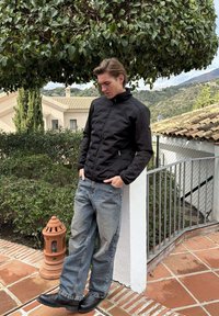 Jonge man in een zwarte jas en wijde spijkerbroek leunt tegen een witte muur buiten, met een gesnoeide boom en een terracotta lantaarn in de buurt.