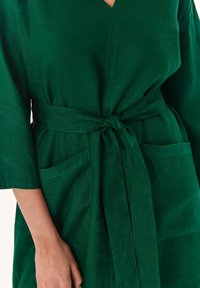 Zielona lniana sukienka kopertowa z dekoltem w kształcie litery V, rękawami o długości trzech kwarty, przewiązywanym w pasie oraz dwiema przednimi kieszeniami. Teksturowany materiał, prosty fason.