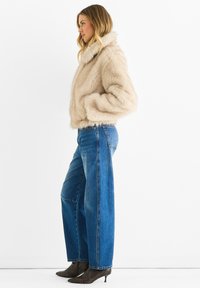 Woman in beige faux fur jacket, wide-leg blue jeans, and pointed brown ankle boots standing sideways on white background.
