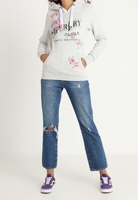 Sudadera gris con detalles florales, presenta un cordón blanco y bolsillos frontales. Combinada con unos vaqueros desgastados de color azul y zapatillas moradas.